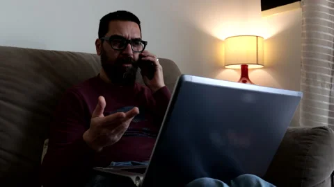 Man sitting on sofa at home working with laptop pc while talking on phone. Stock Footage 239015627