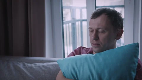 A man is sitting on a sofa, hugging a pillow, with a pensive expression on his Stock Footage 314521560