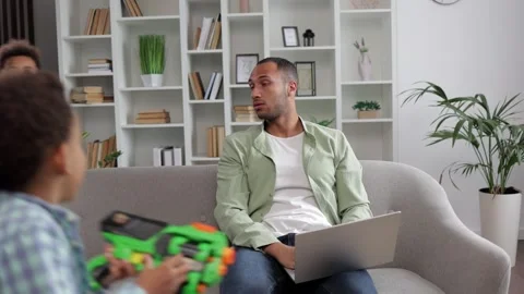 Man sitting on sofa in living room trying to work while two boys of school ag Stock Footage 253587499