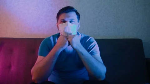 A man sitting on a sofa in a protective mask attentively watches TV with his Stock Footage 131730629