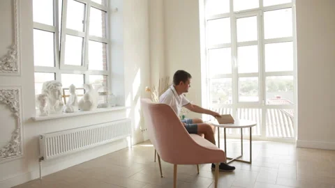 A man is sitting on a sofa in a spacious room, takes a book and begins to read. Stock Footage 205610841