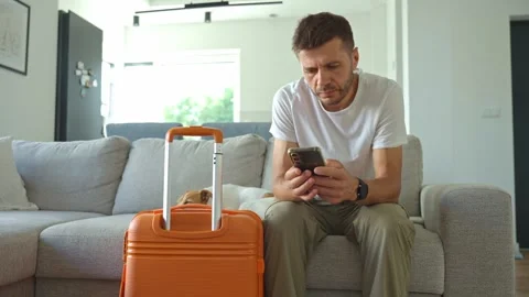 Man sitting on sofa with suitcase and using smartphone Stock Footage 314599328