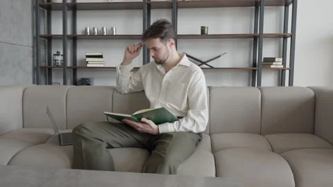 Man Sitting On Sofa, Thinking With Pen In Hand While Reading Notebook Notes Stock Footage 306699594