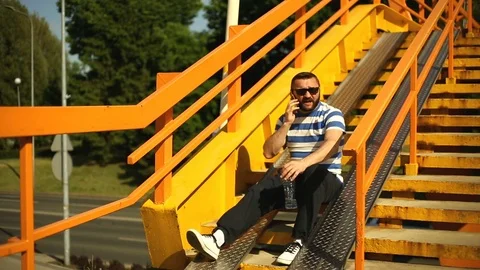 Man sitting on the stairs next to the highway while talking on cellphone Stock Footage