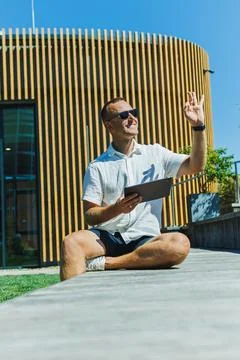 Man sitting on a stone ledge, waving and using a tablet outside a modern bu.. Stock Photos
