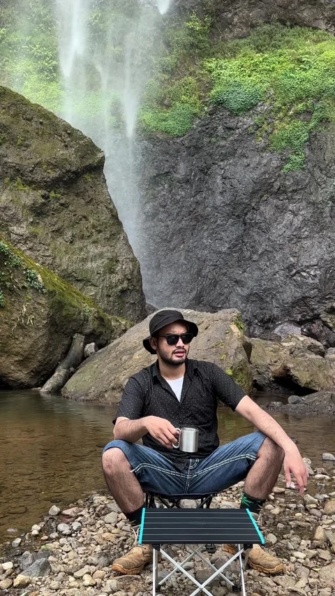 Man Sitting on Stool Drinking by Waterfall in Tropical Forest Vertical Shot Stock Footage 323912267