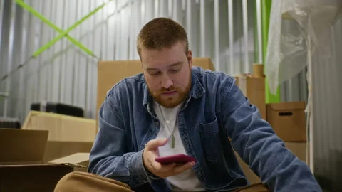 Man Sitting in Storage Unit Using Smartphone and Drinking Coffee Stock Footage 340290876