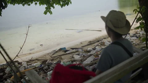 Man sitting on a swing overlooking a polluted tropical beach covered 動画素材 344700504