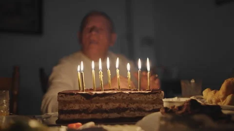A man is sitting at a table blowing the birthday cake that has lit candles on Stock Footage 274481102