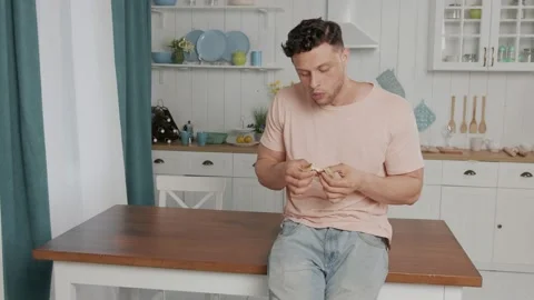Man sitting at the table in a white kitchen and eating a sandwich with sausage Stock-Footage 145451354