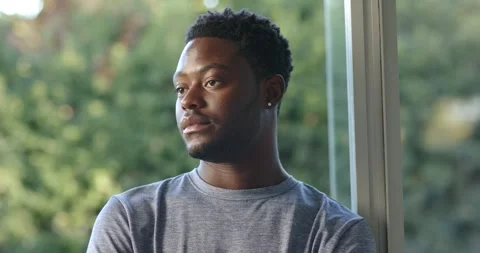 Man sitting by tall window in gray tee, seeing camera, turning, making eye Stock Footage 345037656