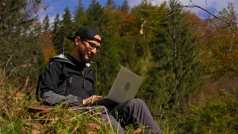 Man sitting on top of a grass covered field. oung man works behind a laptop in Video stock 139871746