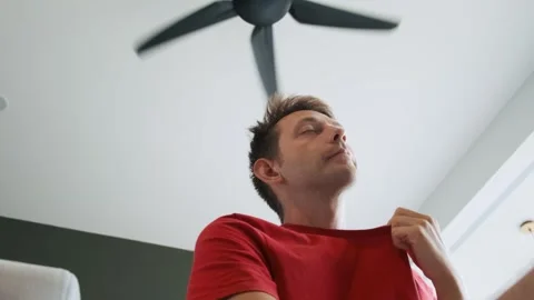 Man sitting under ceiling fan at home in living room, cooling off during hot Stock Footage 278463446