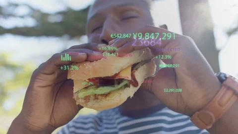 Man sitting under tree eating hamburger, financial charts floating and scaling Stock Footage 332897468
