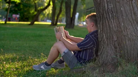 A man is sitting under a tree on the grass in the park and reading a book Stock Footage 156762445