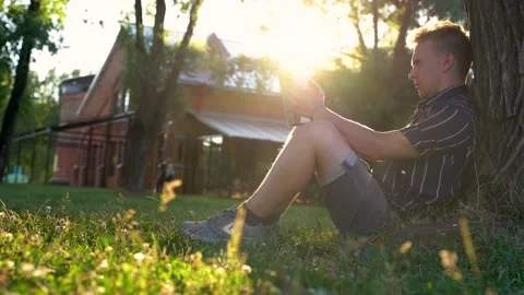 A man is sitting under a tree on the grass in the park and reading a book Stock Footage 156762460