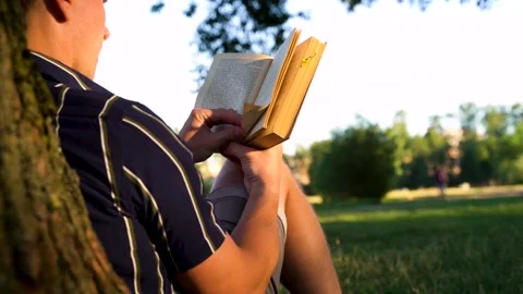 A man is sitting under a tree on the grass in the park and reading a book Stock Footage 156762462