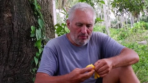A man sitting under the tree while eating a ripe small mango. Video stock 122477043