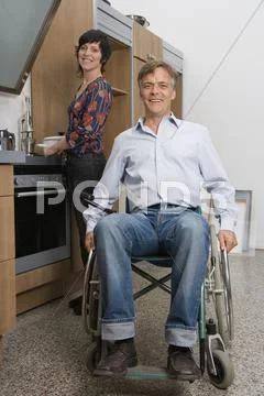 Man sitting in wheelchair and woman standing behind in domestic kitchen Stock Photos