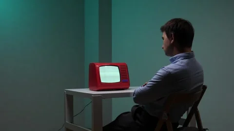 Man sitting at a white table in a dark room, arms crossed, watching a small Stock Footage 345531021
