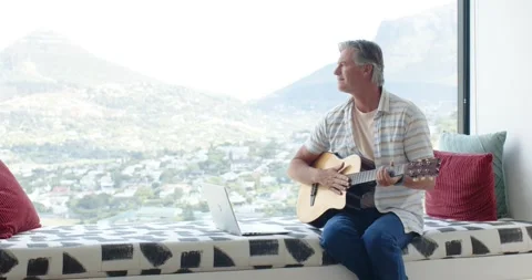 Man sitting by window, glancing out, strumming guitar and checking laptop, copy Stock Footage 335625693