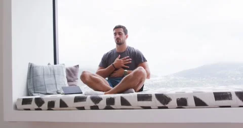 Man sitting on window seat, closing eyes, moving hands to chest, focusing breath Stock Footage 337933330