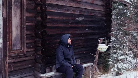 Man Sitting on Wooden Bench Outside Log Cabin During Moderate Snowfall Video stock 334239221