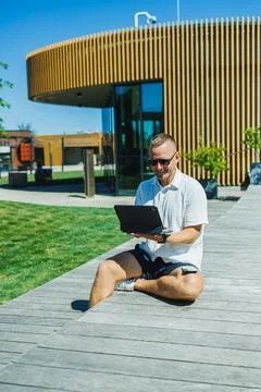 Man sitting on wooden deck using laptop in outdoor area near modern buildin.. Stock Photos