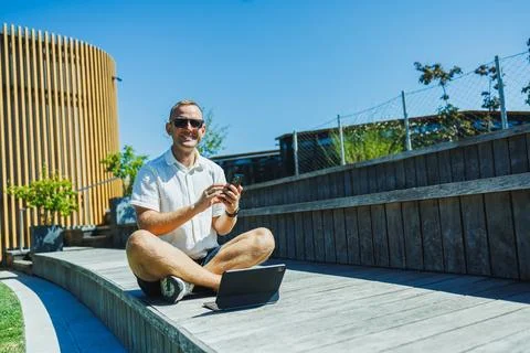 Man sitting on wooden steps using laptop and phone in sunny outdoor setting Stock Photos