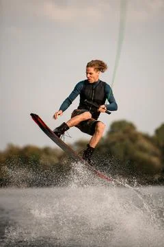 Man skillfully jumping over splashing wave on wakeboard. Sky and green trees in 스톡 사진