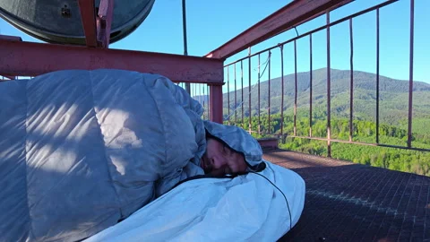 Man in sleeping bag sleeping on 75m tower platform with mountain view Stock Footage 338115651