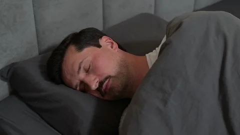 A man is sleeping on a bed covered with a gray blanket. He appears to be in a 動画素材 344527276
