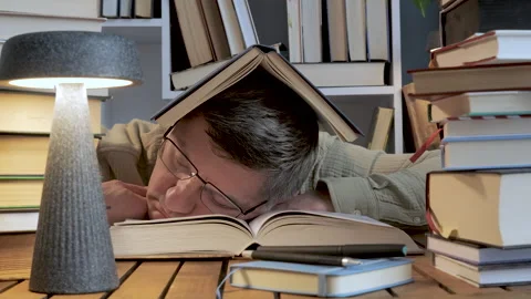 Man sleeping on open book at desk among piles of literature 库存影片 334531869