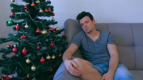 Man is sleeping sitting on couch at home near Christmas tree at eve, hangover. Video stock 136050158