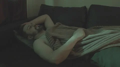 A man sleeps on a sofa under a brown blanket. Concept: mental anxiety and instab Stock Footage 299055671