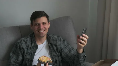 Man with slice of pizza and smartphone looking at camera smiling sitting sofa. Stock Footage 152969720
