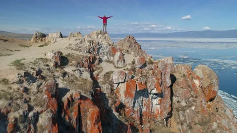 Man in slow motion hands up, stands on cliff top spring lake Baikal ice. Aerial Stock Footage 202644141