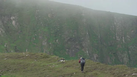 A man with a small backpack walks uphill towards high cliffs on St Kilda Stock Footage 229614892