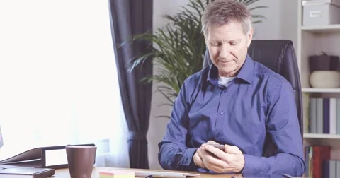 Man with smartphone and typing a message at his desk in a home office 스톡 동영상 61135598