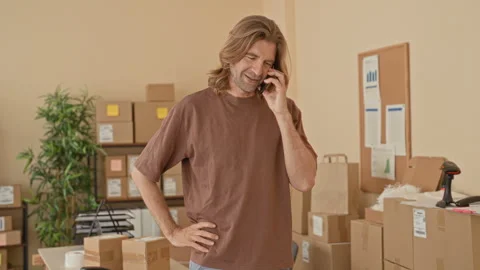 Man with smartphone held to ear, hand on hip beside stacked parcels and car.. Stockbeeldmateriaal 337375931