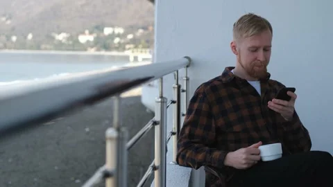 A man with a smartphone sits on a balcony overlooking the sea 스톡 동영상 201051194