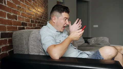 A man with a smartphone sits on a sofa. He is angry and screams Video stock 132647033