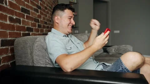 A man with a smartphone sits on a sofa. He is very happy about something Stock Footage 132647376