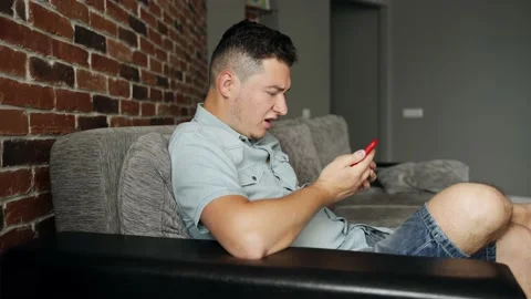 A man with a smartphone sits on a sofa. He is angry and screams Video stock 132647538