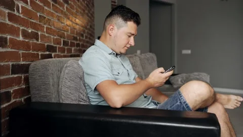 A man with a smartphone sits on a sofa. He has neutral face. He browsing Stock Footage 132647657