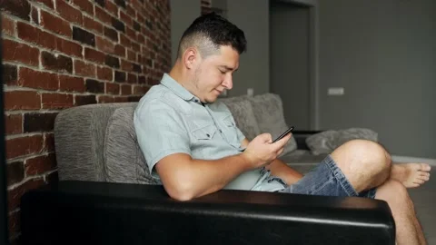 A man with a smartphone sits on a sofa. He has neutral face. He browsing Stock Footage 132647979