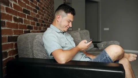 A man with a smartphone sits on a sofa. He has unhappy face. He browsing Stock Footage 132648245
