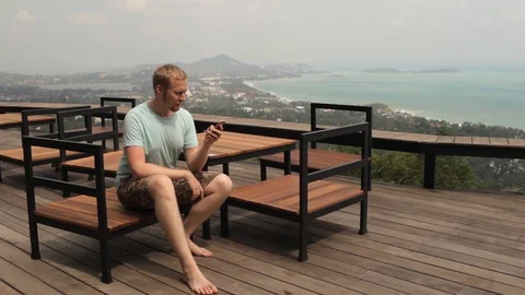 Man with a smartphone is sitting in a cafe overlooking the tropical island Video stock 85934983