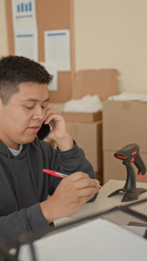Man on smartphone talking while holding a pen at a desk with barcode scanne.. 動画素材 337356065