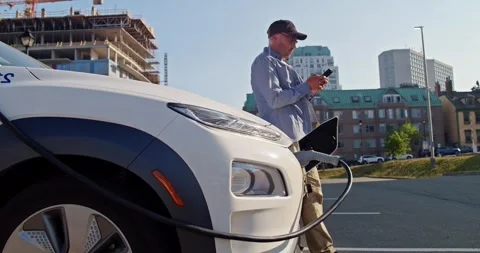 Man with smartphone waits while his electric car charging at a charging station Stock Footage 243326732